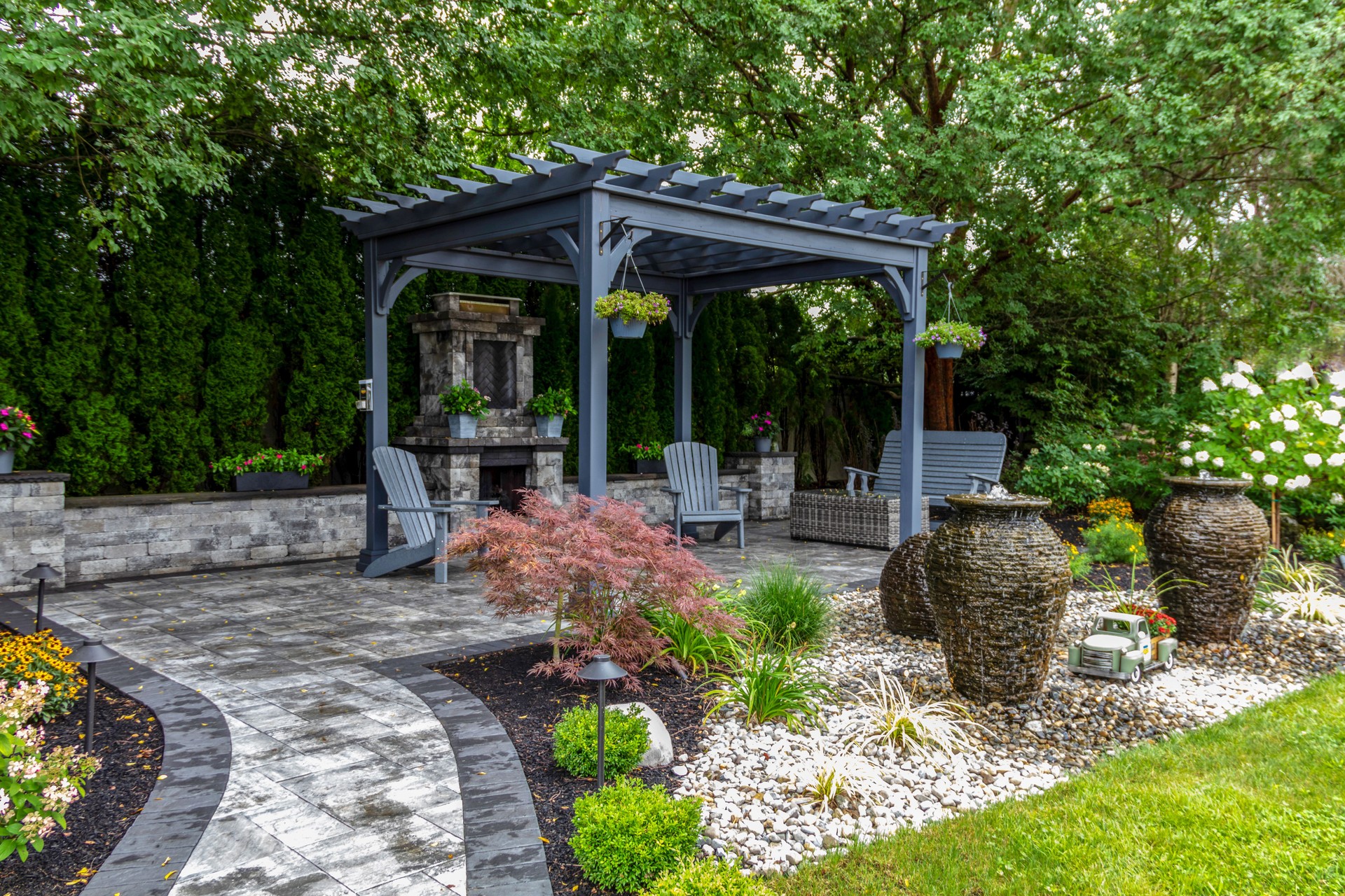 Large gray pergola on pavers with stone fireplace and waterfall urns for backyard oasis landscape design